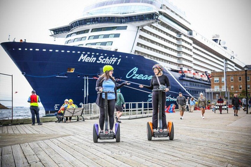 A large cruise ship named 'Mein Schiff 1' docked at a port with people walking and riding Segways on a wooden boardwalk. 1