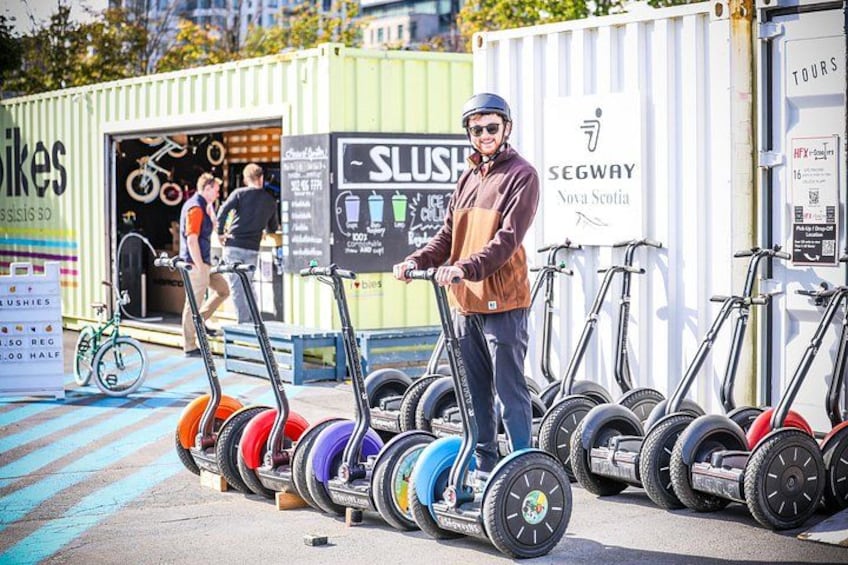 A person standing next to a row of electric scooters, a sign for Segway Nova Scotia, and a SLUSH sign. 2