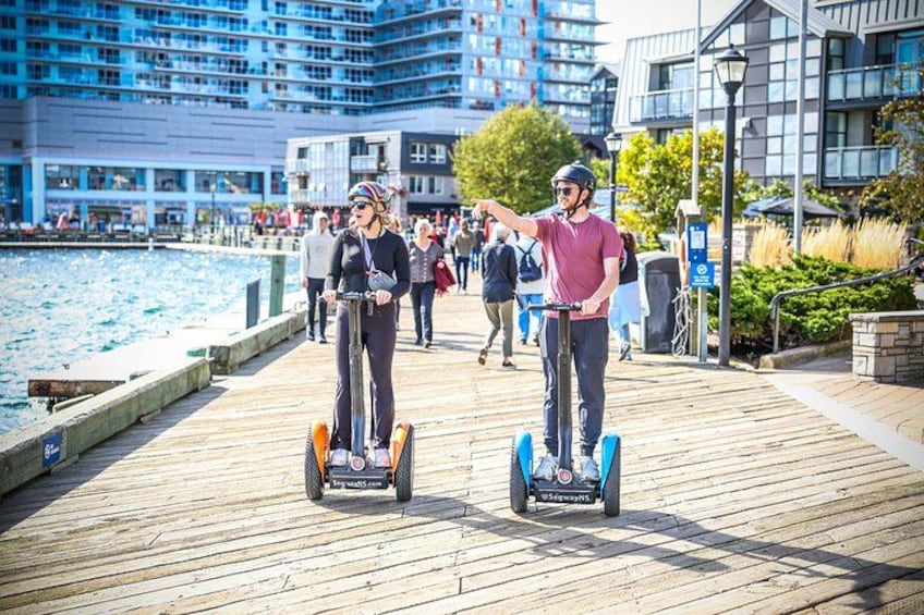 Two people on segways on a wooden boardwalk near a body of water with buildings in the background. 5