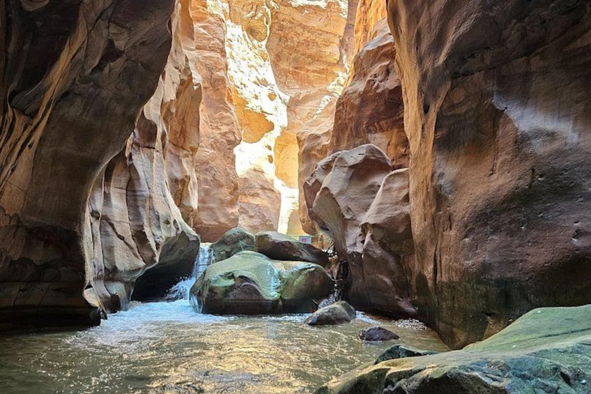 A narrow canyon with a stream flowing through it, surrounded by reddish-brown rock formations. 3