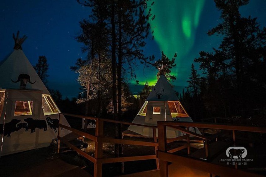 A night scene with two illuminated teepees, a wooden deck, and the aurora borealis. 3