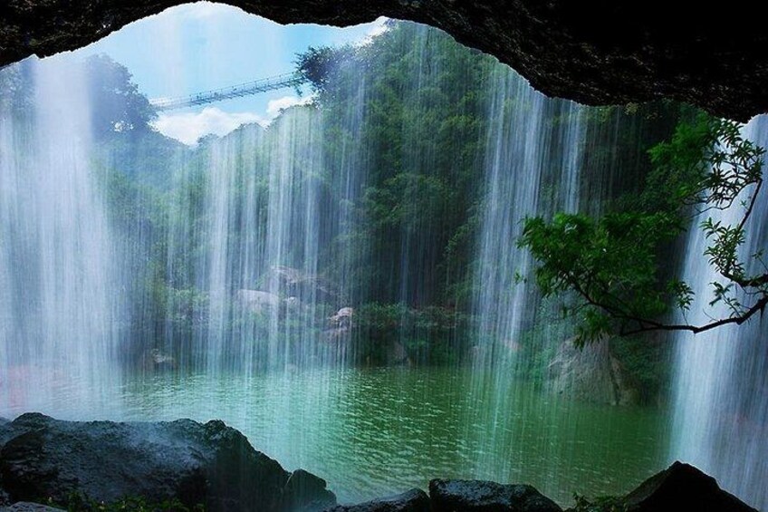 Una cascada se precipita en una piscina tranquila, rodeada de exuberante vegetación y un puente al fondo. 3