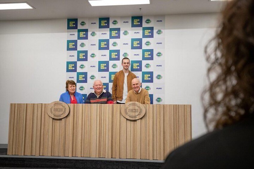 A podium with four people standing behind it, a wall with logos, and a partial view of a person in the foreground. 3
