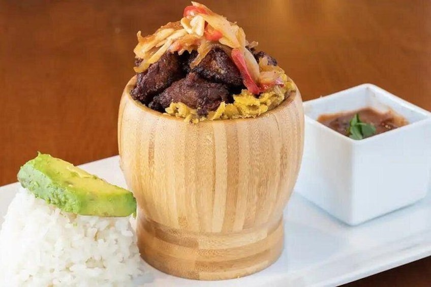 A wooden bowl filled with a meat dish, accompanied by white rice and a slice of avocado, with a small bowl of sauce beside it. 4