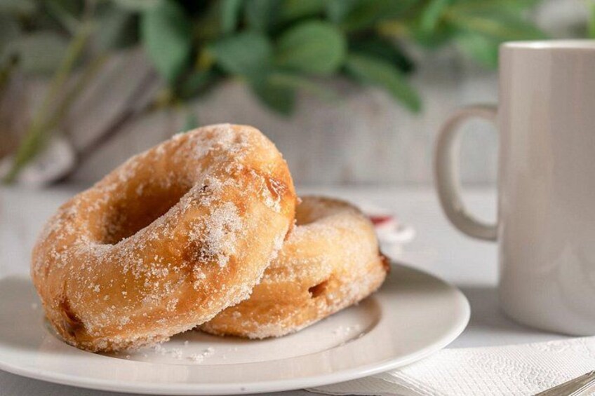 Dos rosquillas cubiertas de azúcar sobre un plato blanco, con una taza blanca al fondo. 3