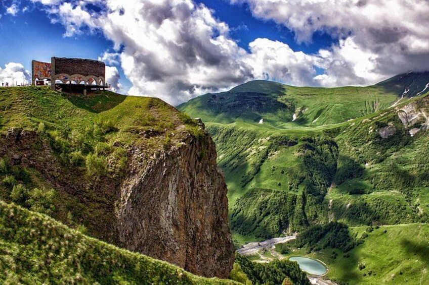Un paisaje montañoso con un edificio en un acantilado, vegetación exuberante y un río. 5