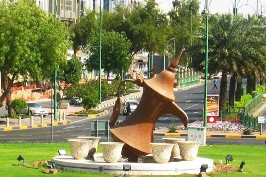 A large abstract sculpture in a public square with surrounding greenery and a busy street. 1