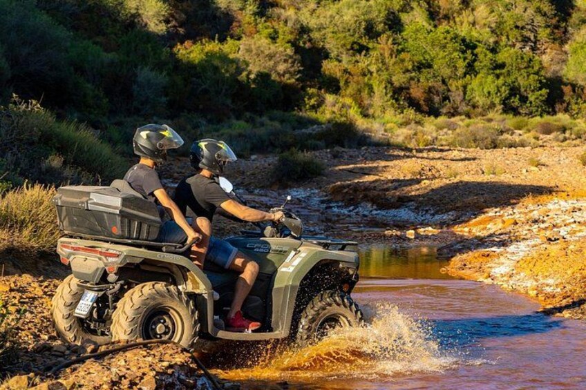 Two people on an ATV crossing a shallow water body in a natural, rocky terrain. 4