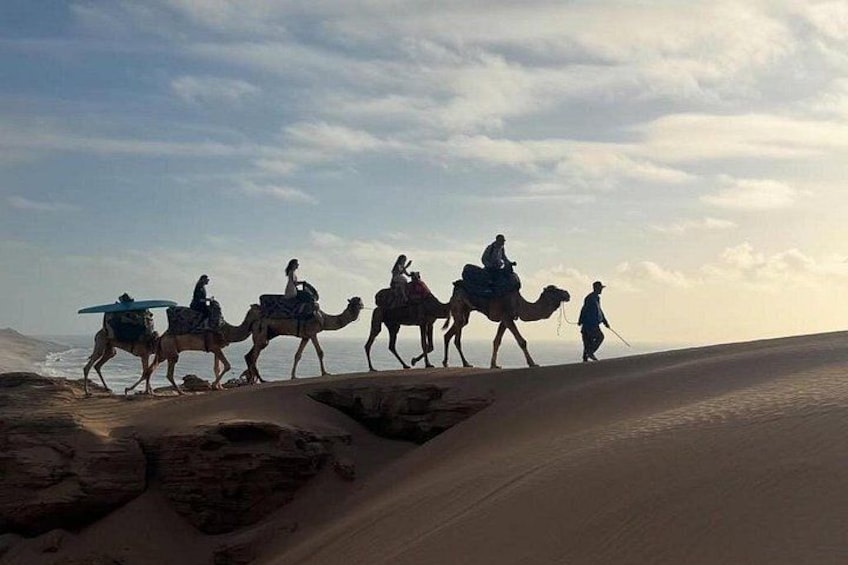 A group of people riding camels across a sandy desert landscape with a body of water in the background. 5
