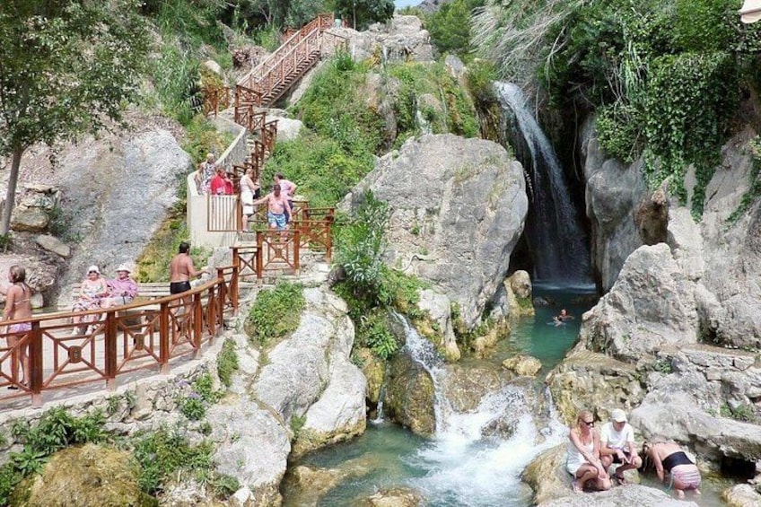 Un manantial termal natural con un paseo de madera, una cascada y personas relajándose. 5