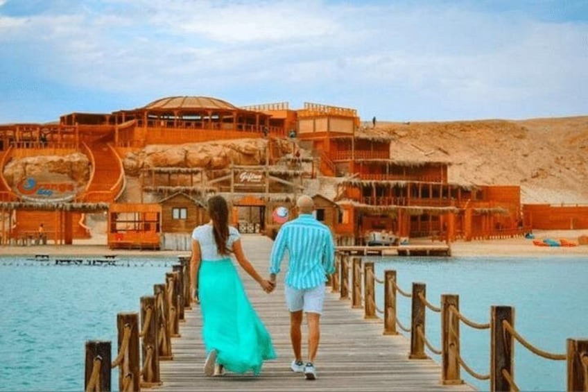 A wooden pier leading to a resort with a pool and a building with a spiral staircase. 4