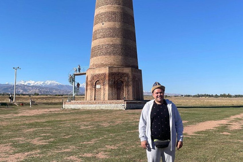 A person standing in front of a tall, cylindrical tower with intricate designs, set against a backdrop of mountains and clear sky. 3