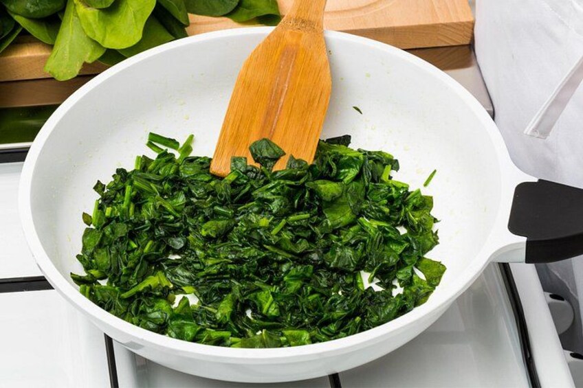 A white pan on a stove with chopped green leafy vegetables being stirred with a wooden spatula. 4