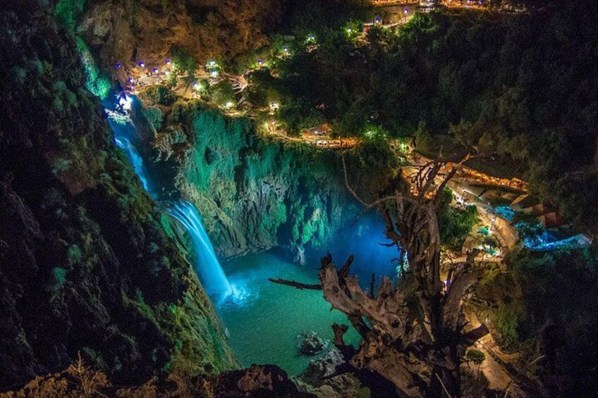 Vista nocturna de un cañón con cascada, senderos iluminados y un pequeño poblado. 4