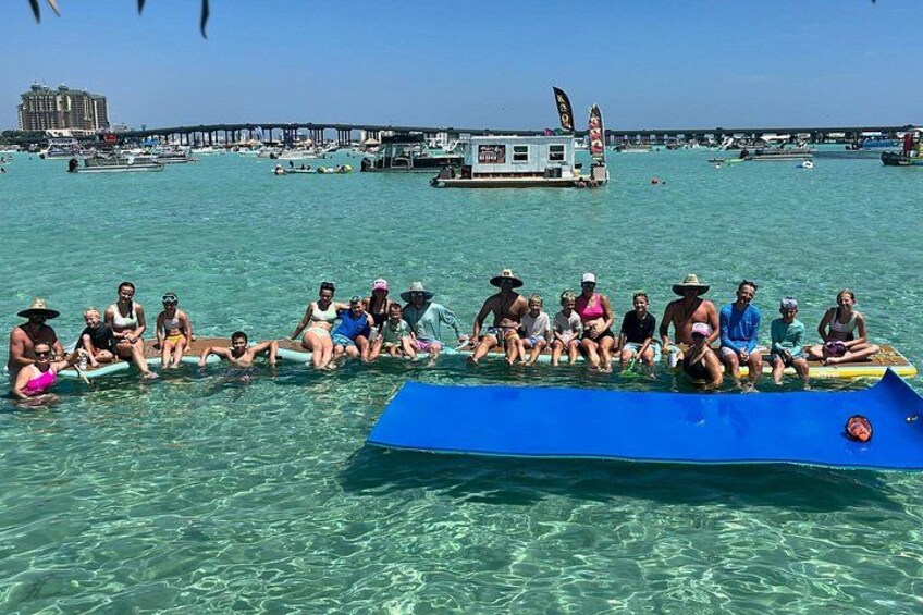 A group of people sitting on a blue raft in clear water with a pier and boats in the background. 5
