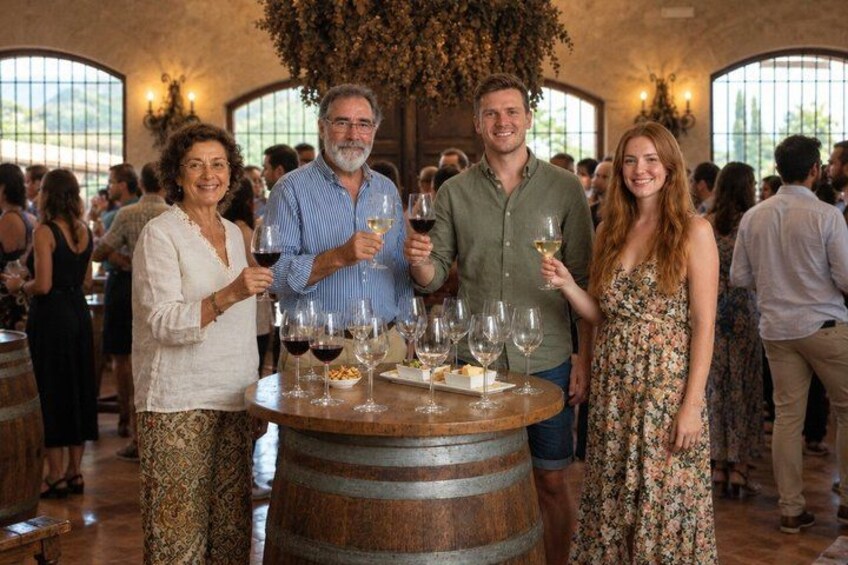 A group of people standing around a round wooden table with wine glasses and bottles. 2