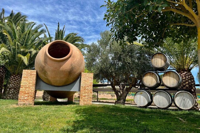 Una gran tinaja de barro y barriles de madera apilados en un área cubierta de pasto, con palmeras y un muro de ladrillo. 1