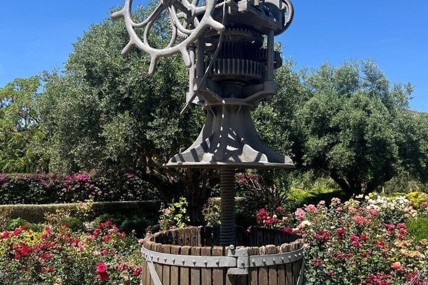 Una escultura con un diseño intrincado, rodeada por una base de piedra circular, ubicada en un jardín con plantas en flor. 5