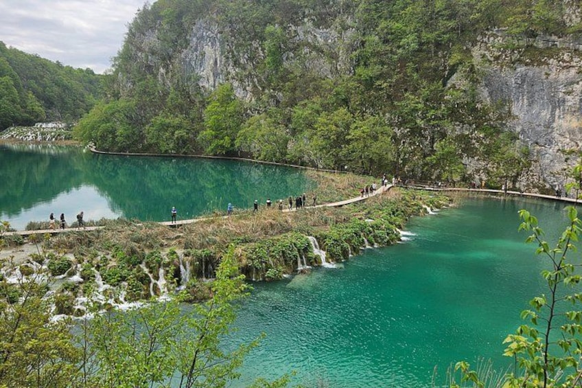 A turquoise lake surrounded by cliffs and greenery, with a walking path and people enjoying the scenery. 1