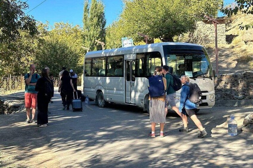 Een groep mensen stapt op een bus, met bomen en een stenen muur op de achtergrond. 1