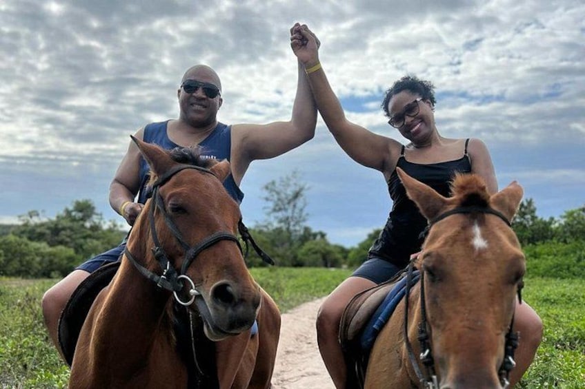 Two people riding horses on a dirt path with greenery in the background. 2