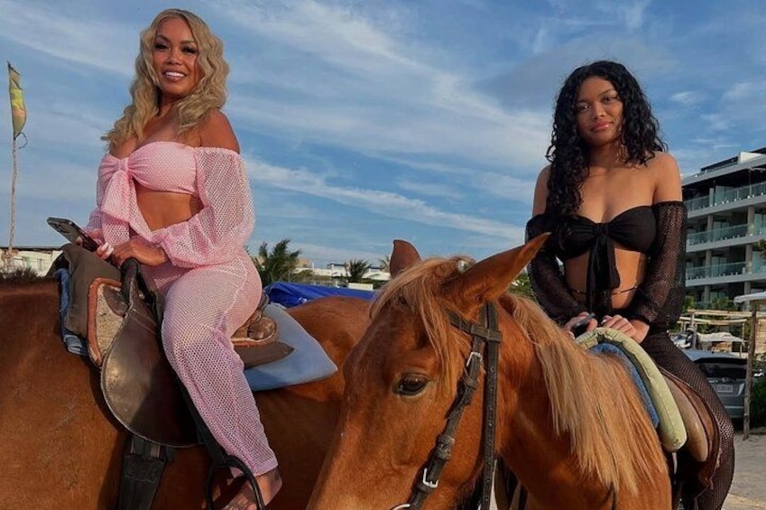 Zwei Personen reiten auf Pferden am Strand, im Hintergrund sind Gebäude zu sehen. 2