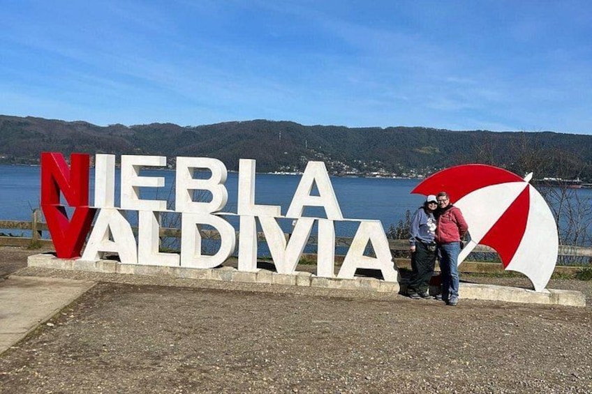 Een bord met de tekst 'NIEBLIA VALDIVIA' en een meer met bergen op de achtergrond. 1