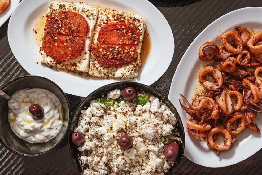 A table with various dishes including grilled octopus, tomato slices on bread, and a bowl of feta cheese with olives. 5