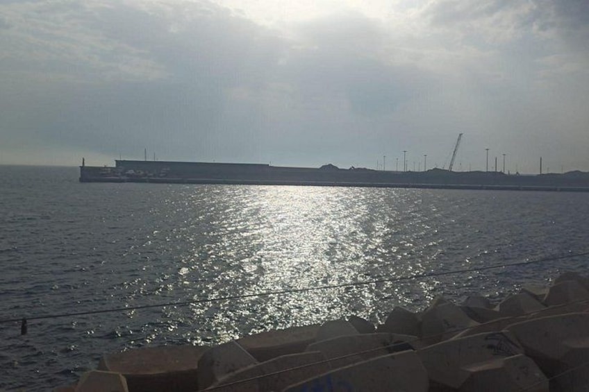 A calm sea with a large ship in the distance, a breakwater, and industrial structures on the horizon. 5