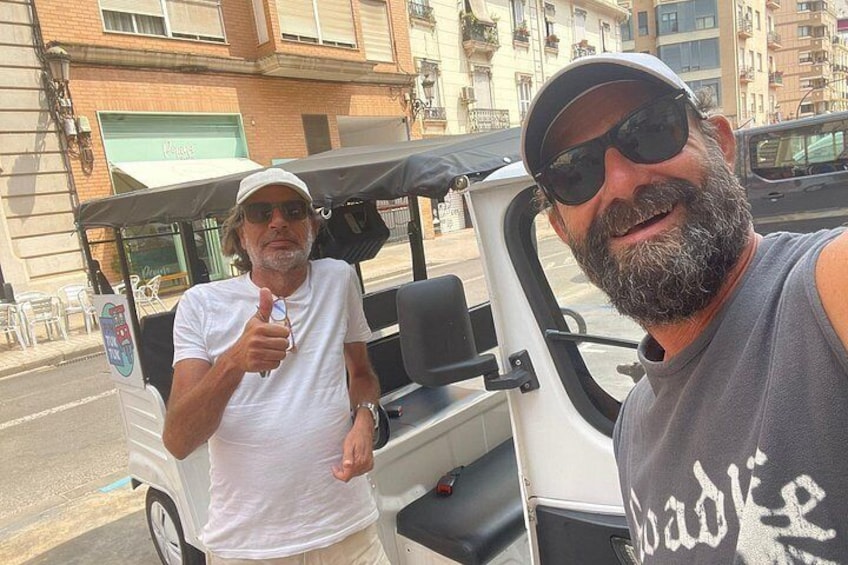 Dos hombres de pie junto a un carrito de golf en una calle con edificios al fondo. 4
