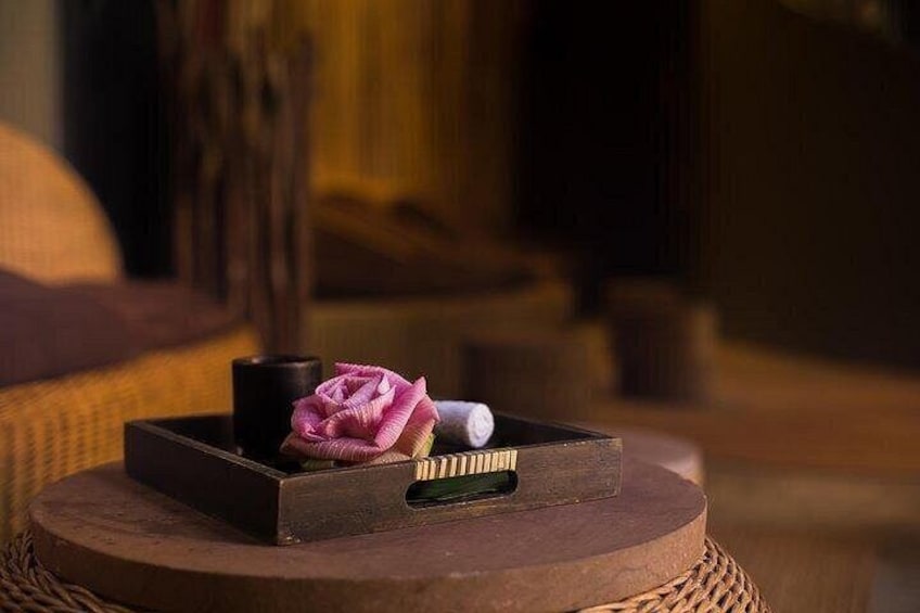 Una bandeja de madera con una flor rosa, una vela y un objeto blanco sobre una mesa redonda de mimbre. 4