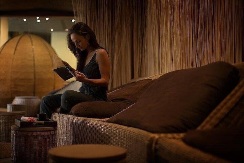 Una persona sentada en un sofá marrón en una habitación con poca luz, con un objeto redondo de madera grande al fondo. 2