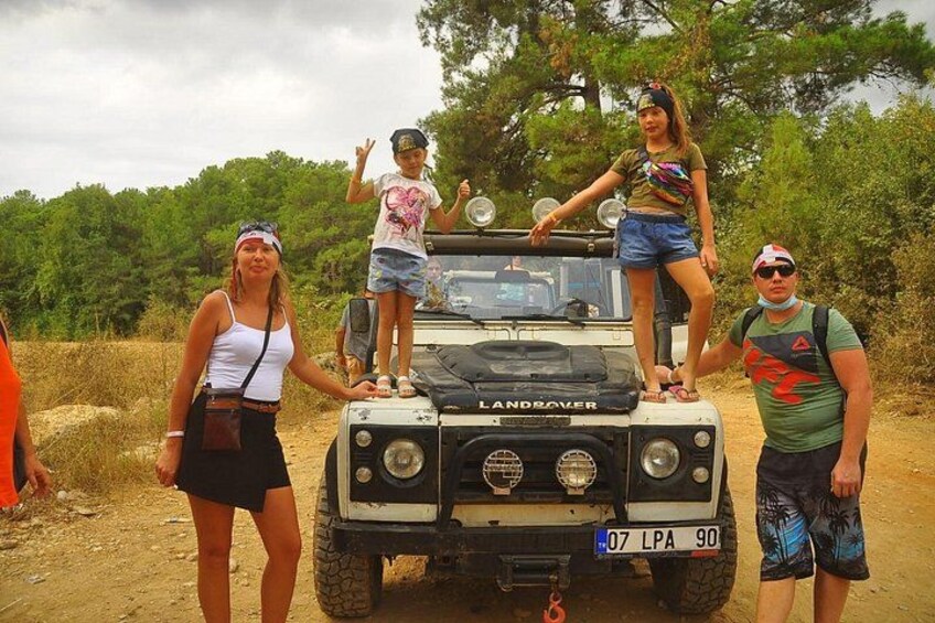 A Land Rover with three people standing on it, a dirt road, and trees in the background. 4