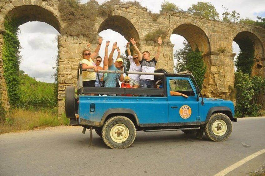 A blue off-road vehicle with passengers waving, parked in front of stone arches. 2
