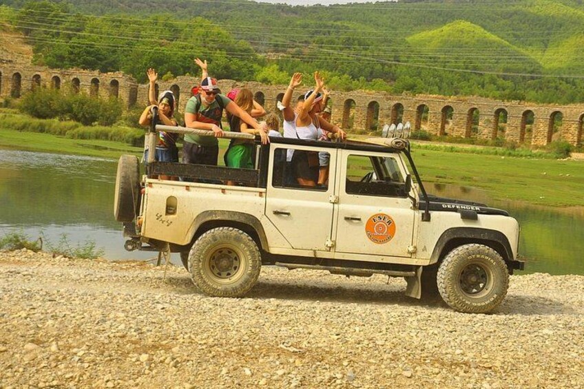 A beige off-road vehicle with passengers waving, a river, and a stone bridge in the background. 1