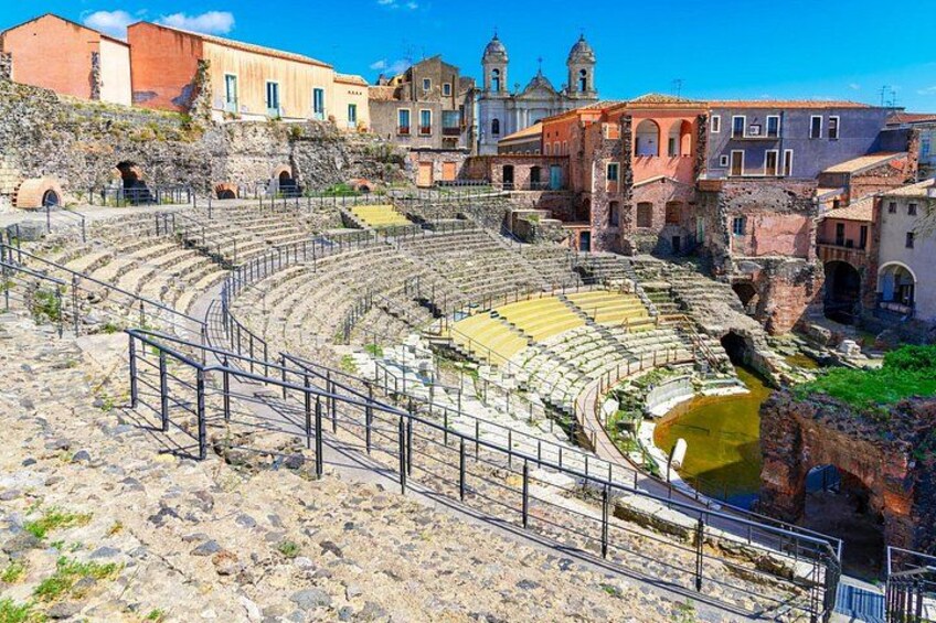 An ancient amphitheater with stone seating and a central stage, surrounded by historic buildings. 2
