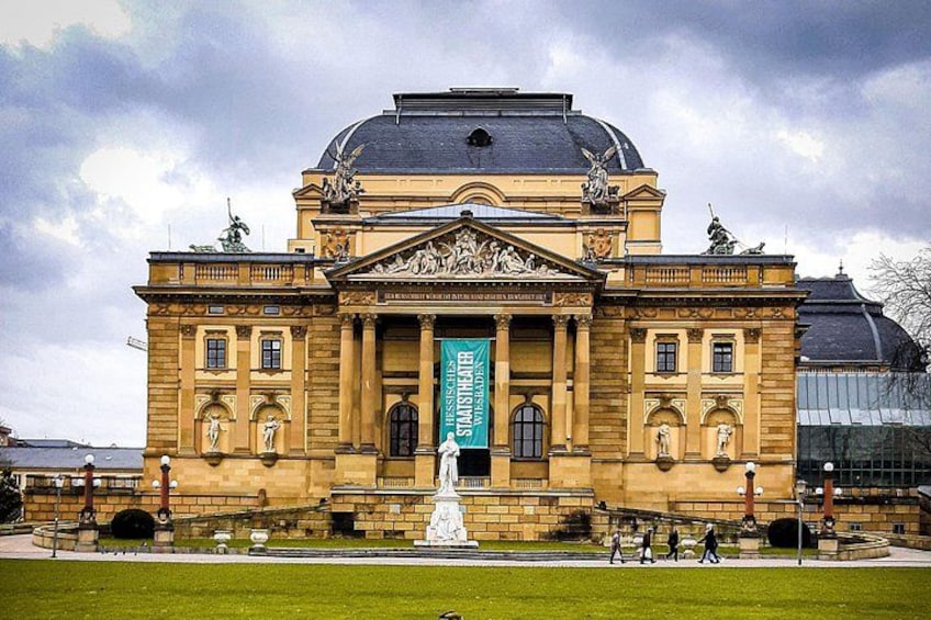 Un bâtiment de style classique, doté d’un dôme central et de statues, entouré d’une pelouse bien entretenue et d’une fontaine. 4