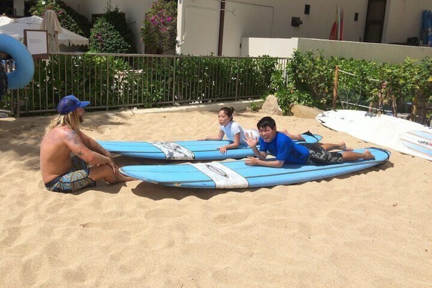 Tre personer ligger på surfboards på en sandstrand med et hegn og vegetation i baggrunden. 3