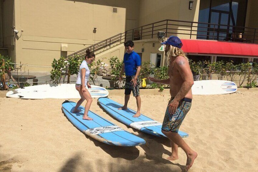 Personer øver sig på surfboards på en sandstrand med en bygning i baggrunden. 2