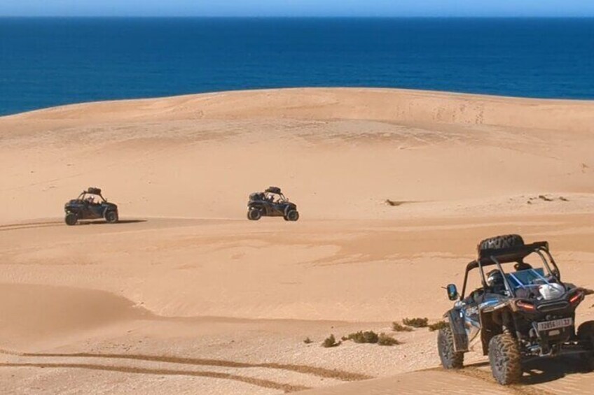 Three off-road vehicles on a desert sand dune with the ocean in the background. 1