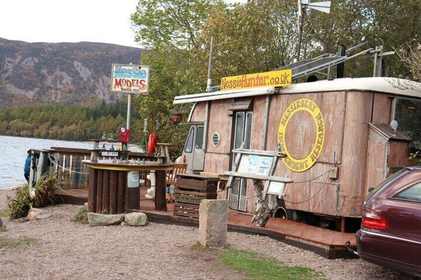 Ein hölzernes Boot mit einem Schild, auf dem 'Nessie Hunter' und 'Modelle seit 1971' steht, am Ufer eines Sees. 2