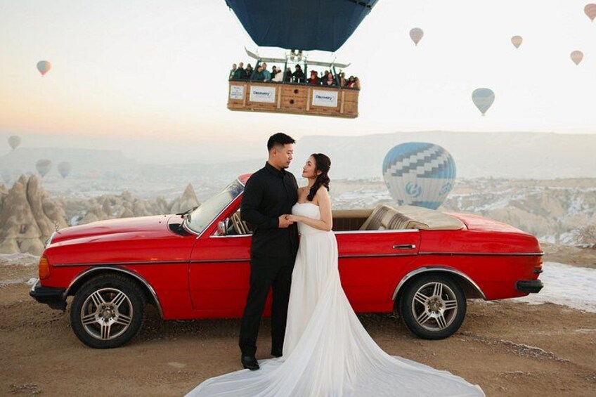 A couple standing next to a red convertible car with a hot air balloon in the background. 1