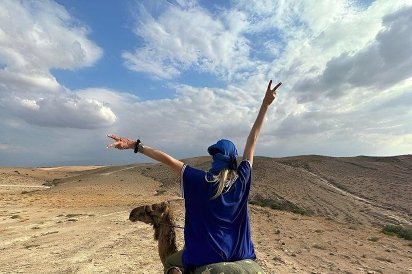 A person in a blue shirt and cap with arms raised, standing on a desert terrain with a camel nearby. 3