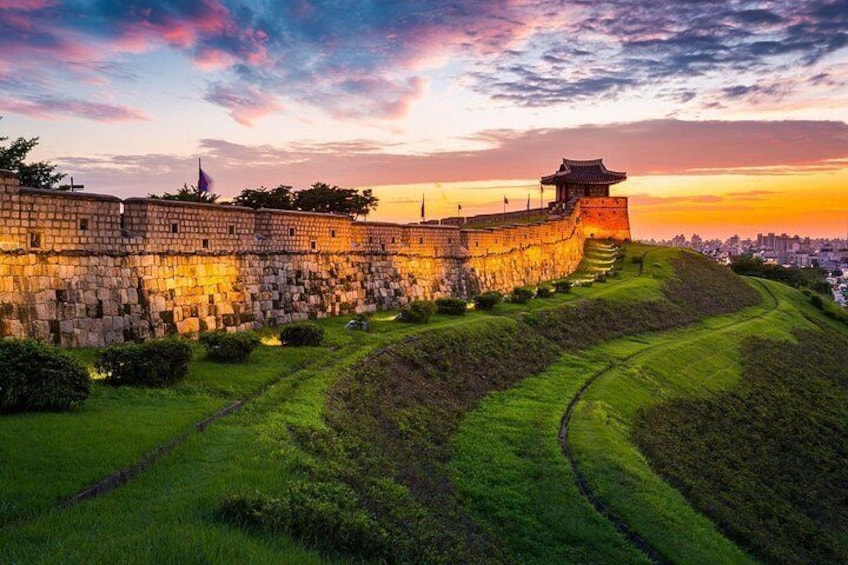A historic stone wall with a traditional roof structure, surrounded by green grass and a cityscape in the background. 1