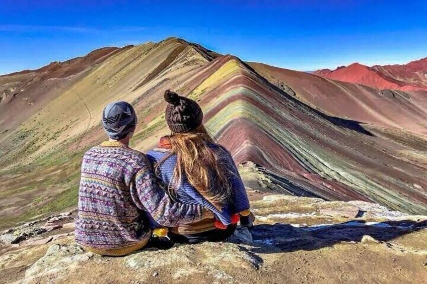 Two people sitting on a rocky outcrop overlooking a colorful mountain range. 1