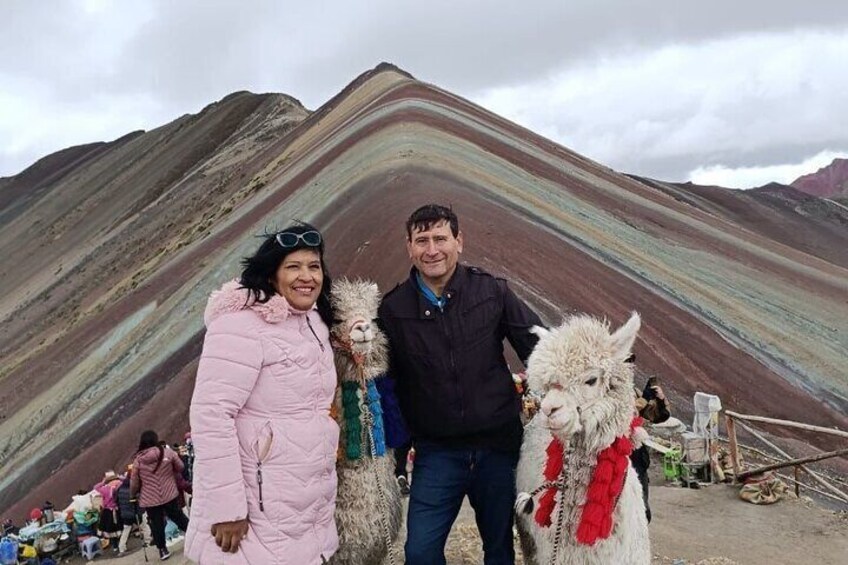 A person in a pink coat and another in a dark jacket standing in front of a colorful mountain range with llamas. 5