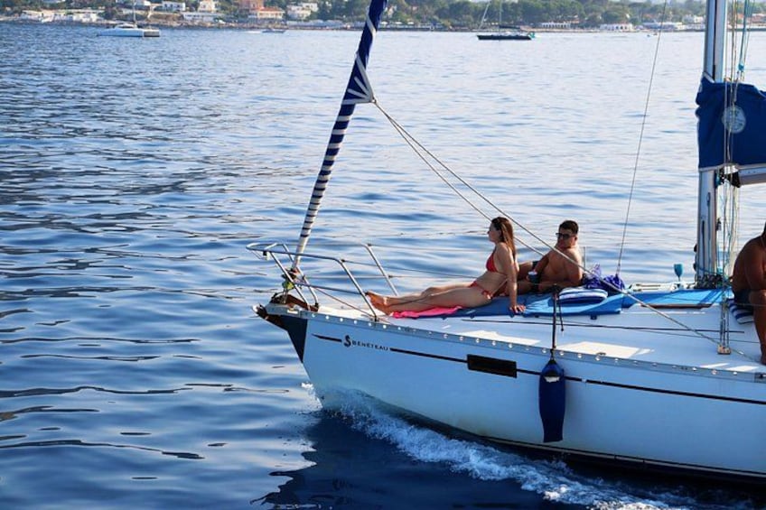 A sailboat with people on board, a clear blue sea, and distant buildings on the shore. 2