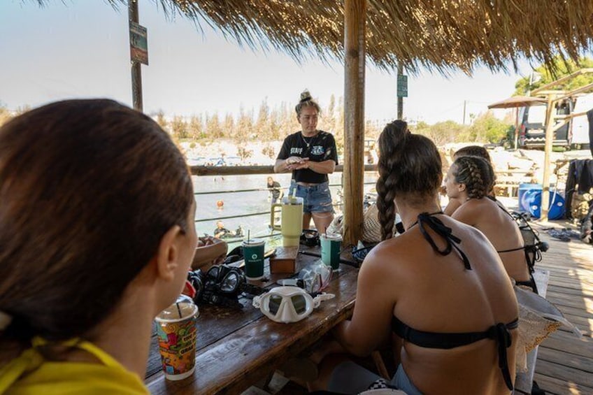 Eine Gruppe von Menschen sitzt an einem Holztisch am Strand, während eine Person steht und mit ihnen spricht. 3