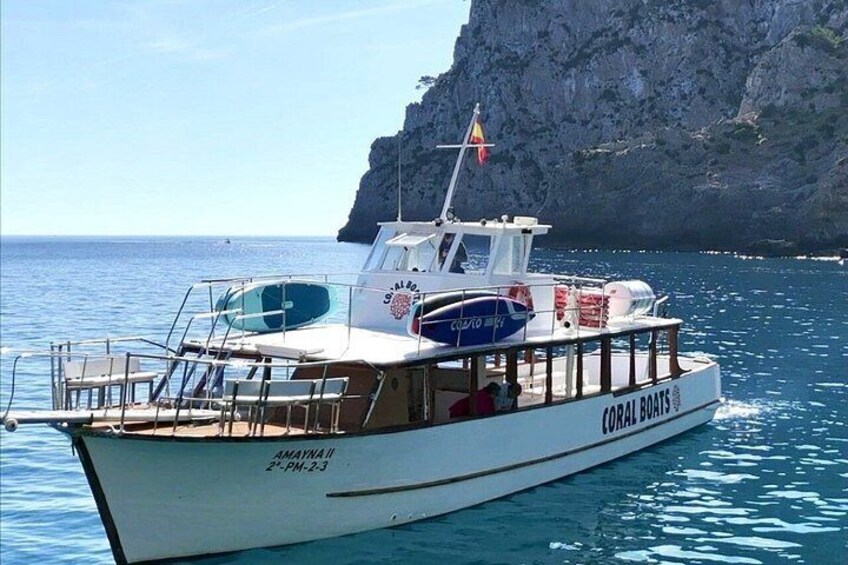 Een boot genaamd 'Coral Boats' met een vlag, afgemeerd nabij een rotsachtige kustlijn. 2