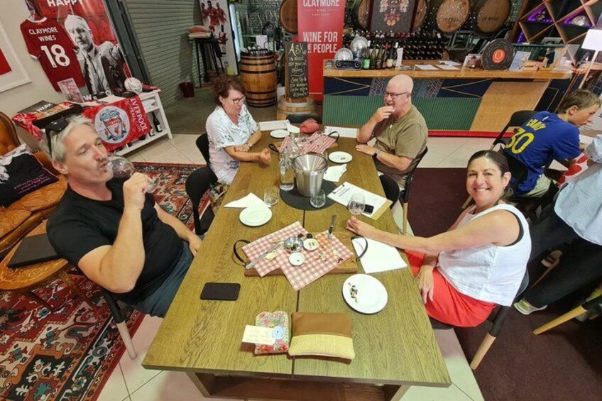 A group of people sitting around a wooden table in a restaurant with a red banner and a sign that reads 'CLAYMORE WINE FOR THE PEOPLE'. 5